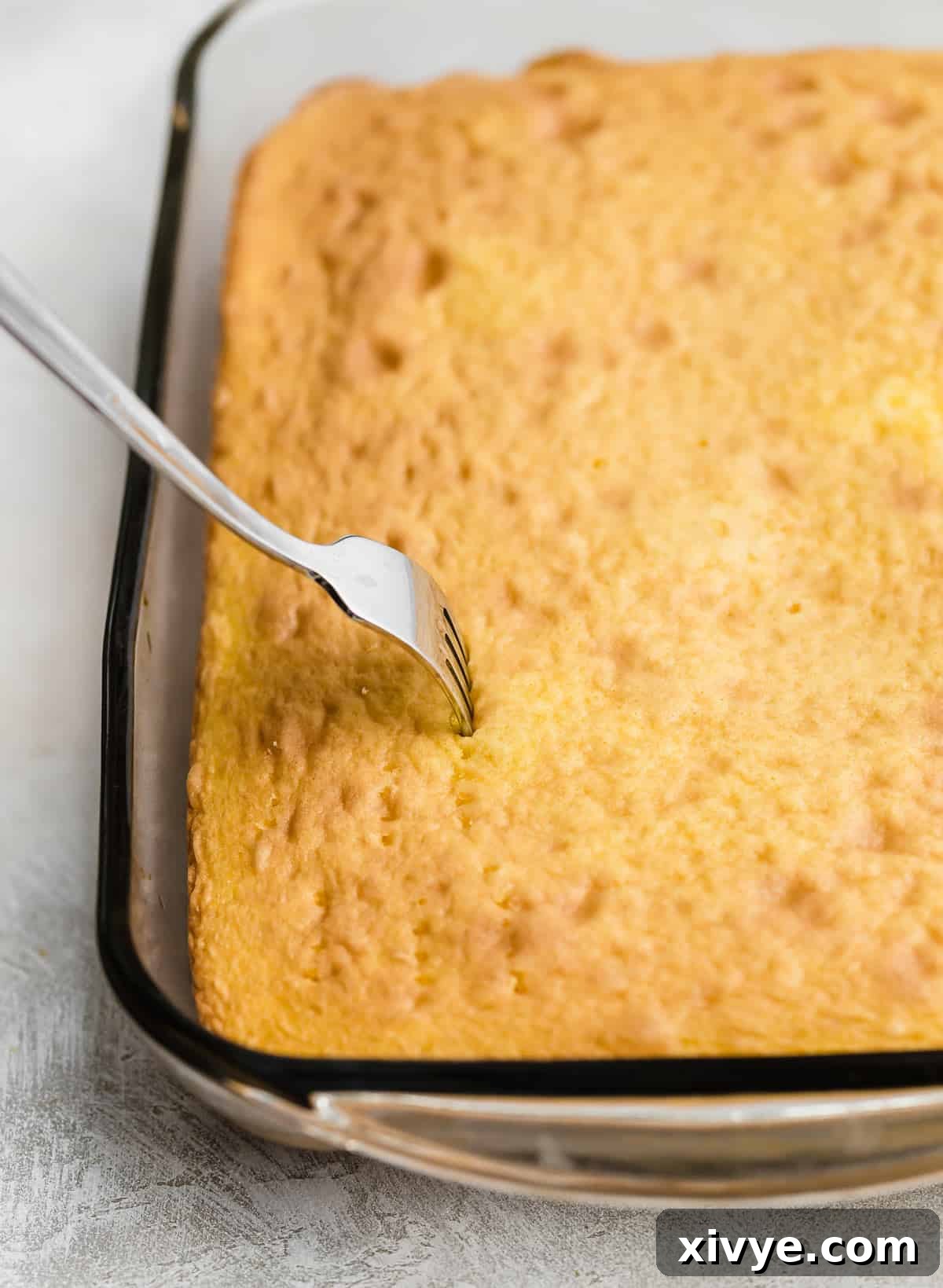 A fork meticulously poking numerous holes across the surface of a baked lemon cake, preparing it for the Jell-O infusion.