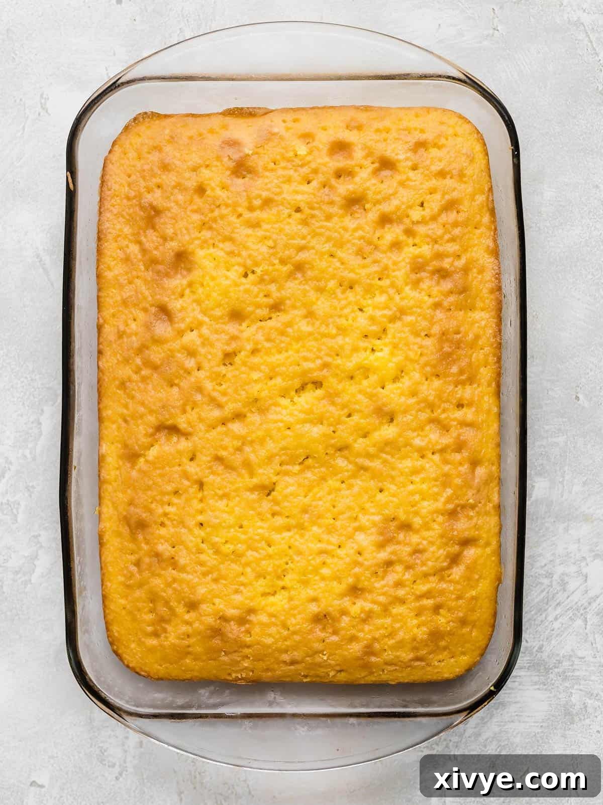 A golden-baked lemon cake resting in a rectangular glass pan, awaiting its transformation.