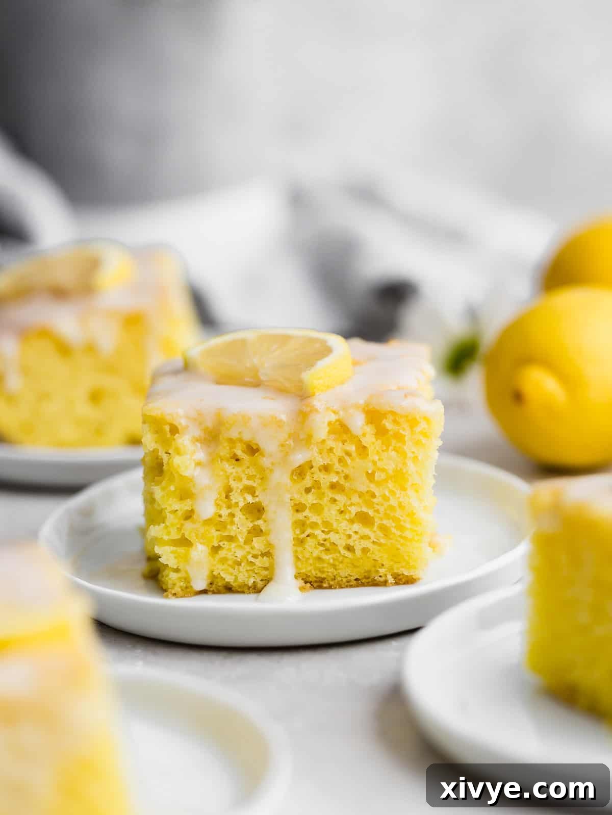 A delicious slice of Lemon Jello Cake, glistening with white glaze and crowned with a vibrant fresh lemon wedge, sits invitingly on a white plate.