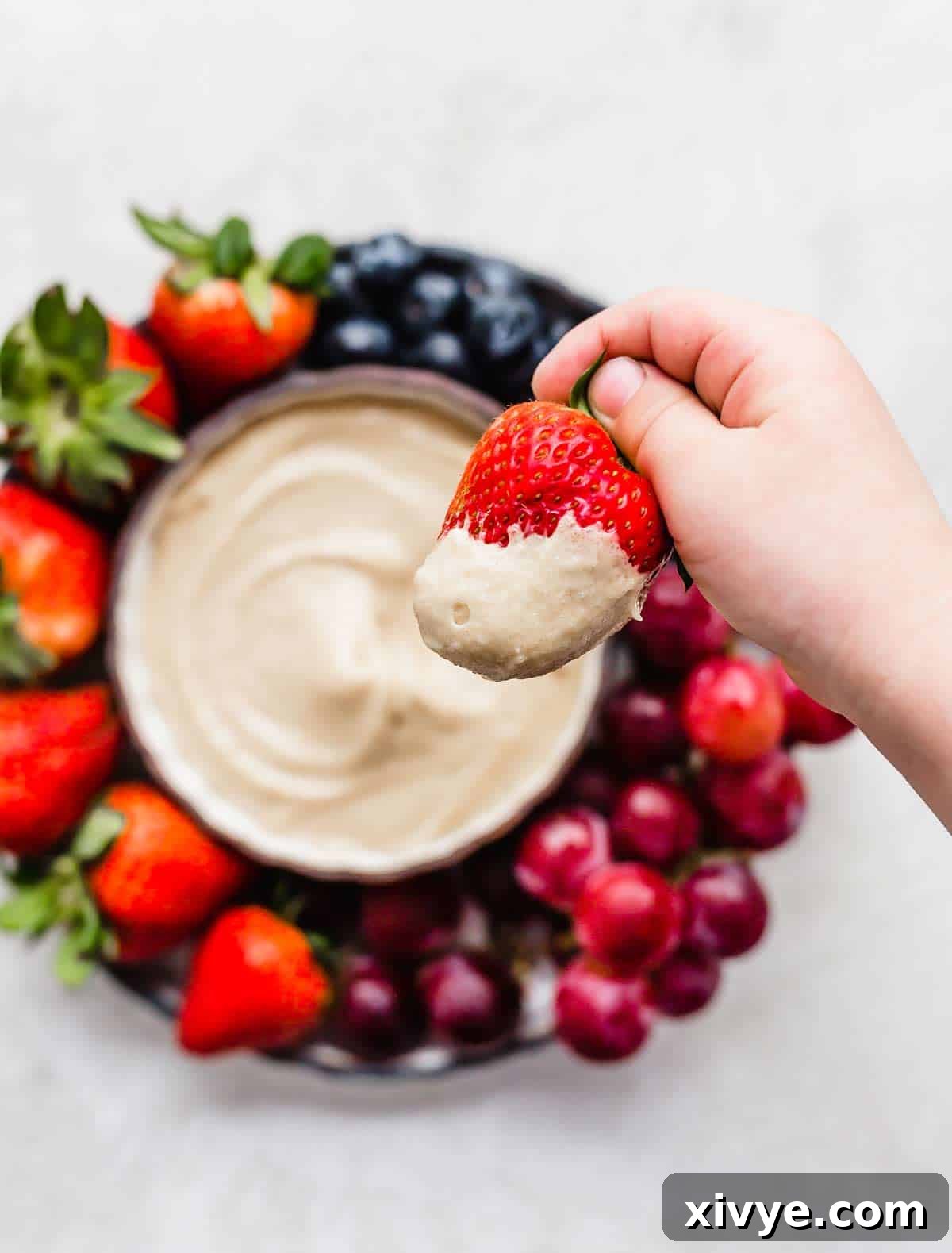 A hand dipping a strawberry into a tan colored Cream Cheese Fruit Dip.