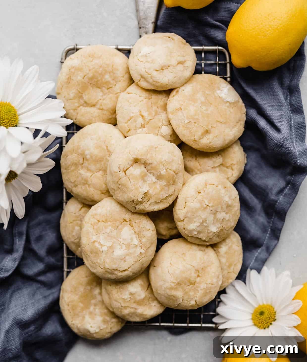 A stack of Lemon Cookies on a small wire rack that is resting on a dark blue linen napkin with two daisy flowers near the cookies.