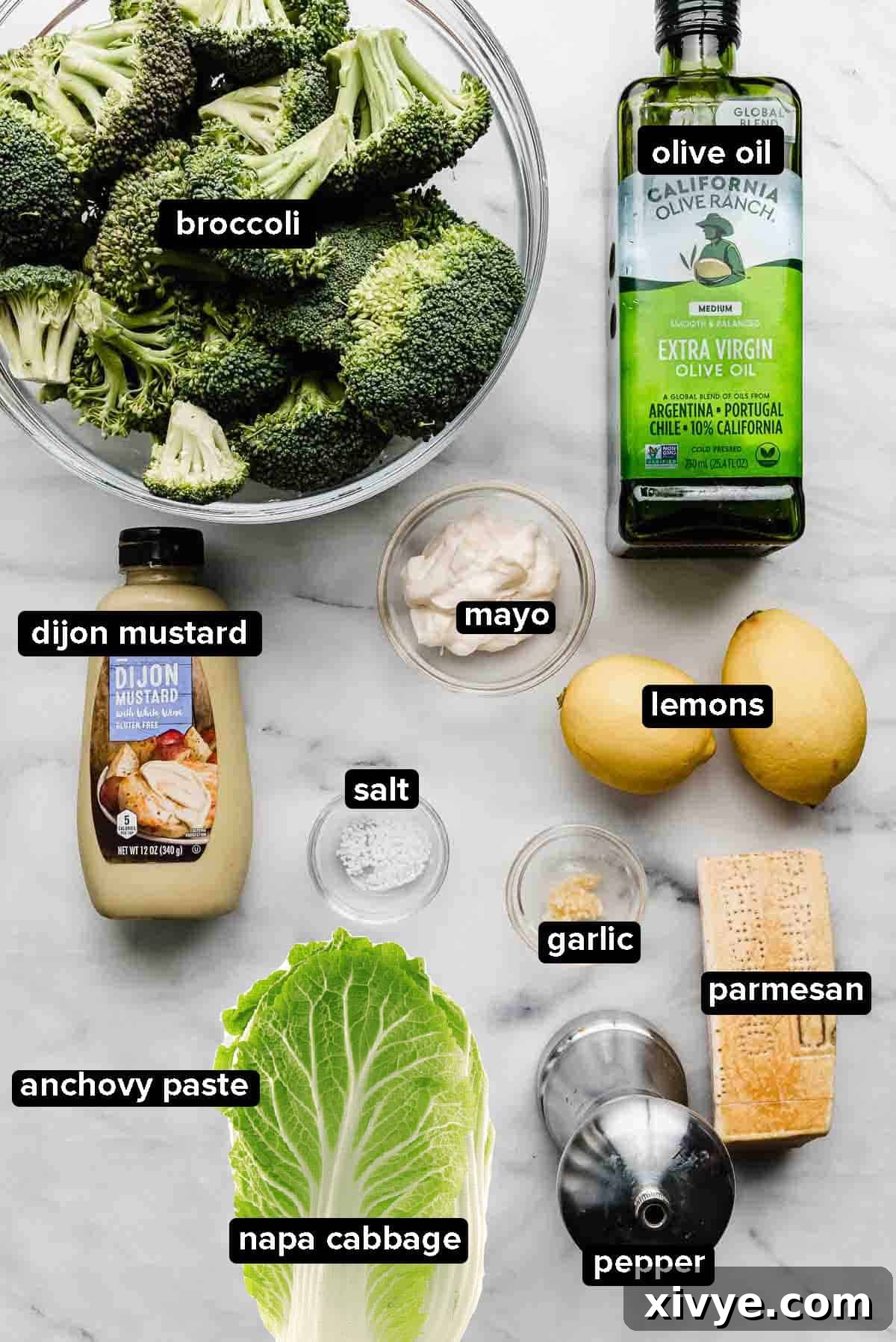Various ingredients for Broccoli Caesar Salad and Napa cabbage meticulously arranged on a white and gray marble backdrop, featuring fresh broccoli, a wedge of Parmesan, whole lemons, a bottle of olive oil, and anchovy paste.