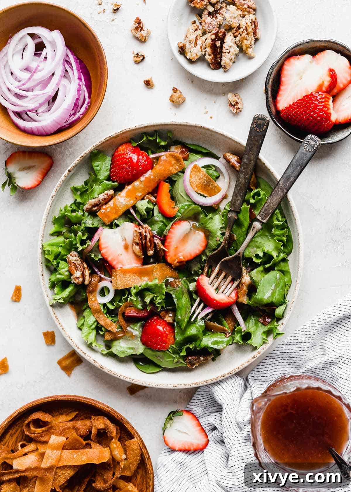 A bowl filled with vibrant Strawberry Spinach Salad, artfully garnished and surrounded by individual bowls of purple onion, fresh strawberries, crispy fried wontons, and glistening candied pecans. The scene exudes freshness and culinary appeal.