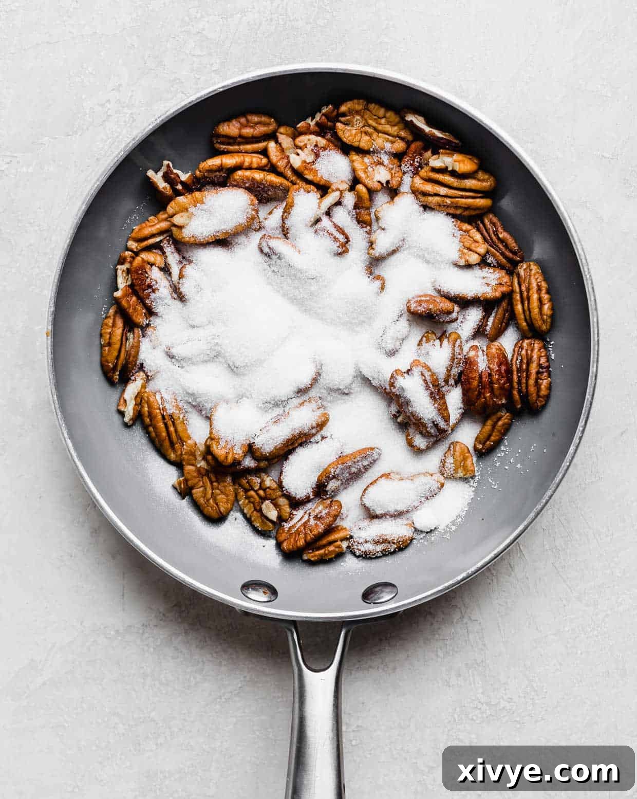 Whole pecan halves arranged in a gray skillet, with granulated sugar sprinkled over them, illustrating the initial stage of the candying process.