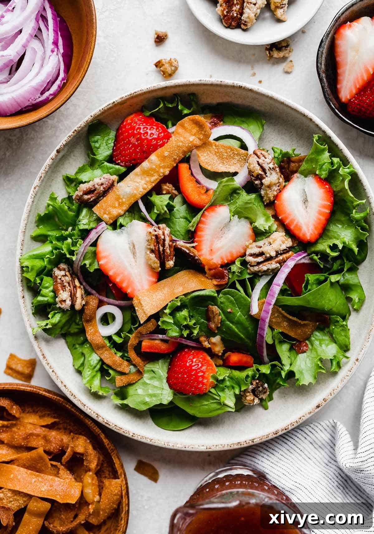 A crisp white plate adorned with a fresh bed of spinach and mixed greens, artfully topped with thinly sliced strawberries, crispy fried wontons, and delicate purple onions, awaiting the final drizzle of dressing.