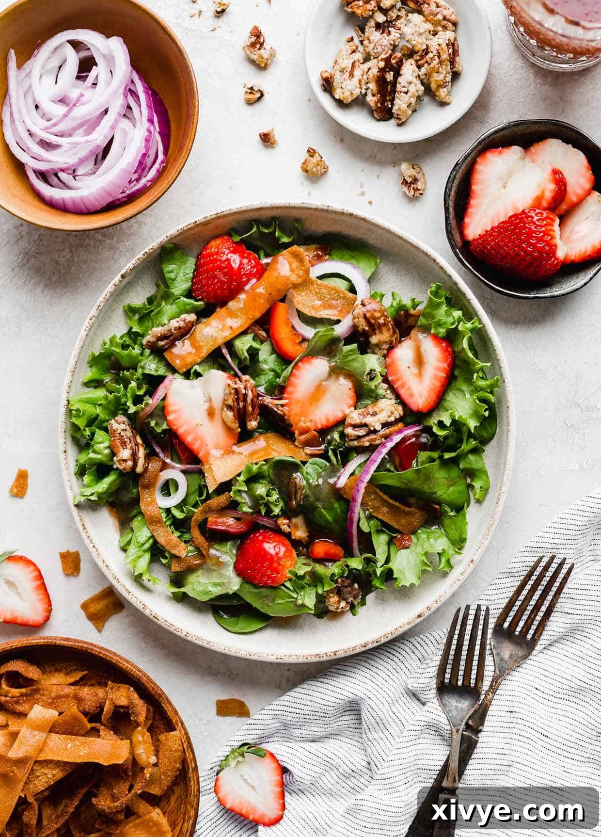 An inviting overhead photo of a vibrant strawberry spinach salad presented on a pristine white background. The salad is artfully surrounded by two forks, scattered fresh strawberries, crispy fried wontons, and thinly sliced red onions, highlighting its beautiful and appetizing presentation.