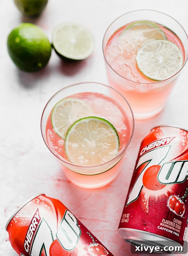 A glass of limeade slush with fresh limes and a can of Cherry 7-UP surrounding it on a rustic surface.