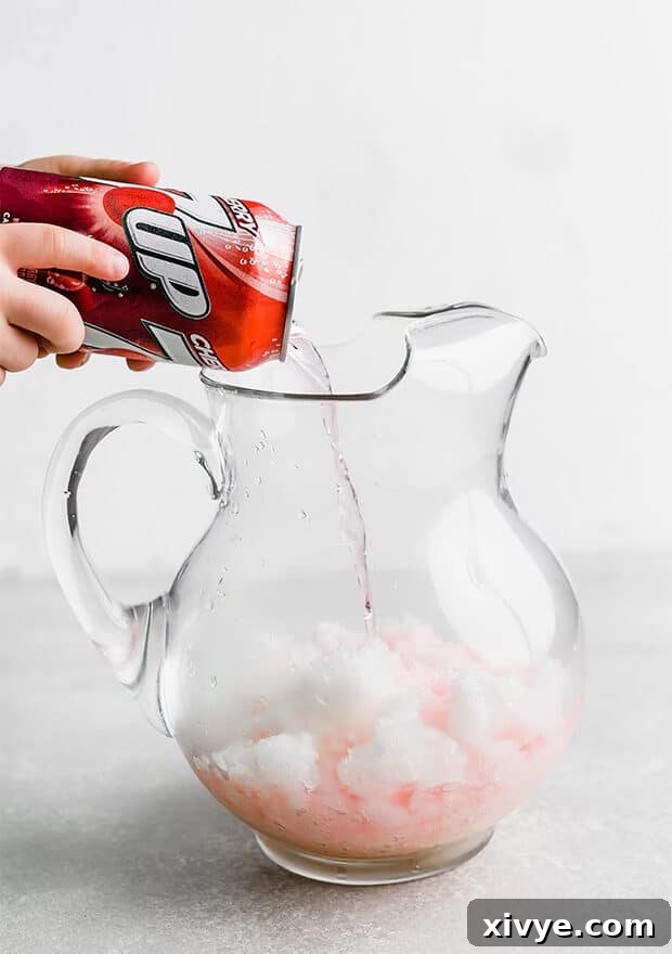 A stream of Cherry 7-UP being poured into a clear glass pitcher filled with green limeade slush.