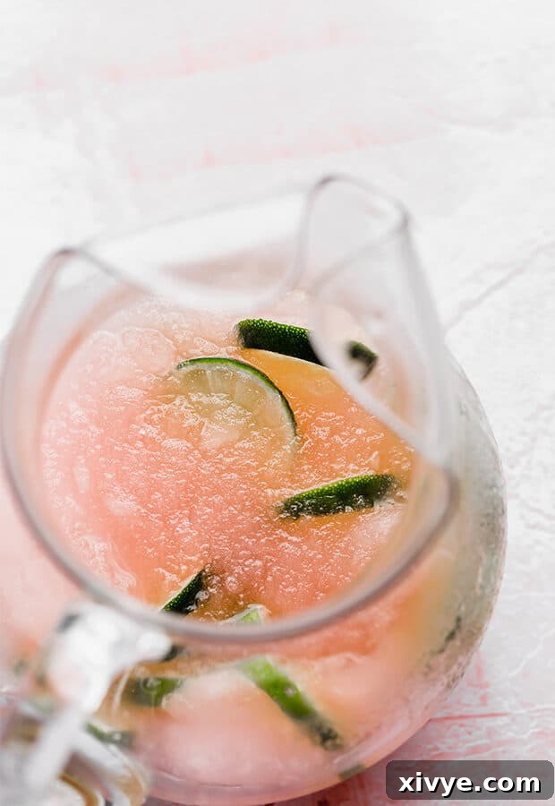 Overhead photo of a clear glass pitcher filled with pink limeade slush, garnished with vibrant green lime slices.