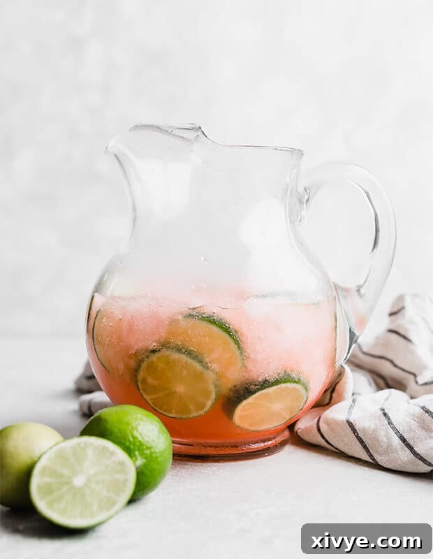 A clear glass pitcher full of vibrant green Limeade Slush Punch, adorned with fresh lime slices, ready to be served.