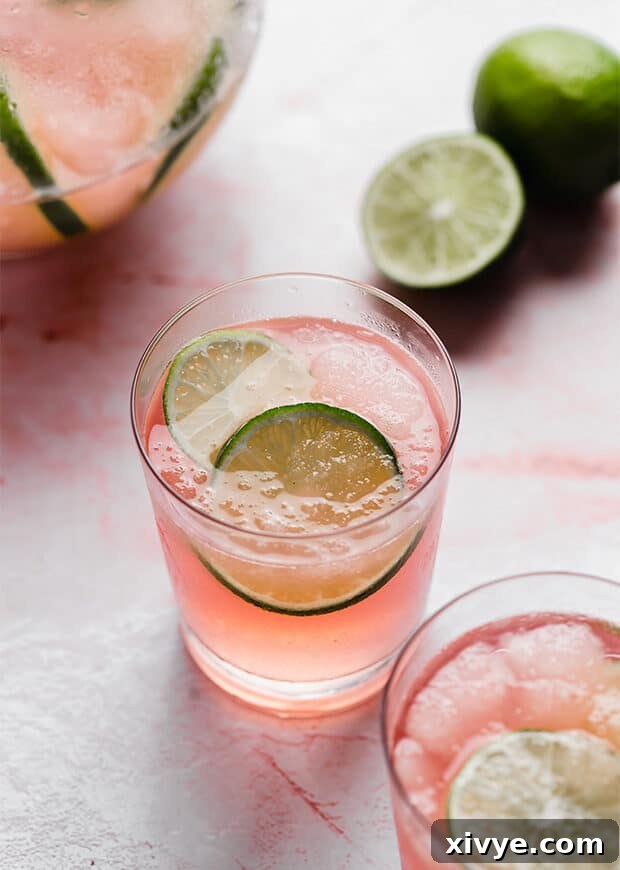 A close-up of a single glass of Limeade Slush Punch, with two lime slices nestled within the icy pink drink.