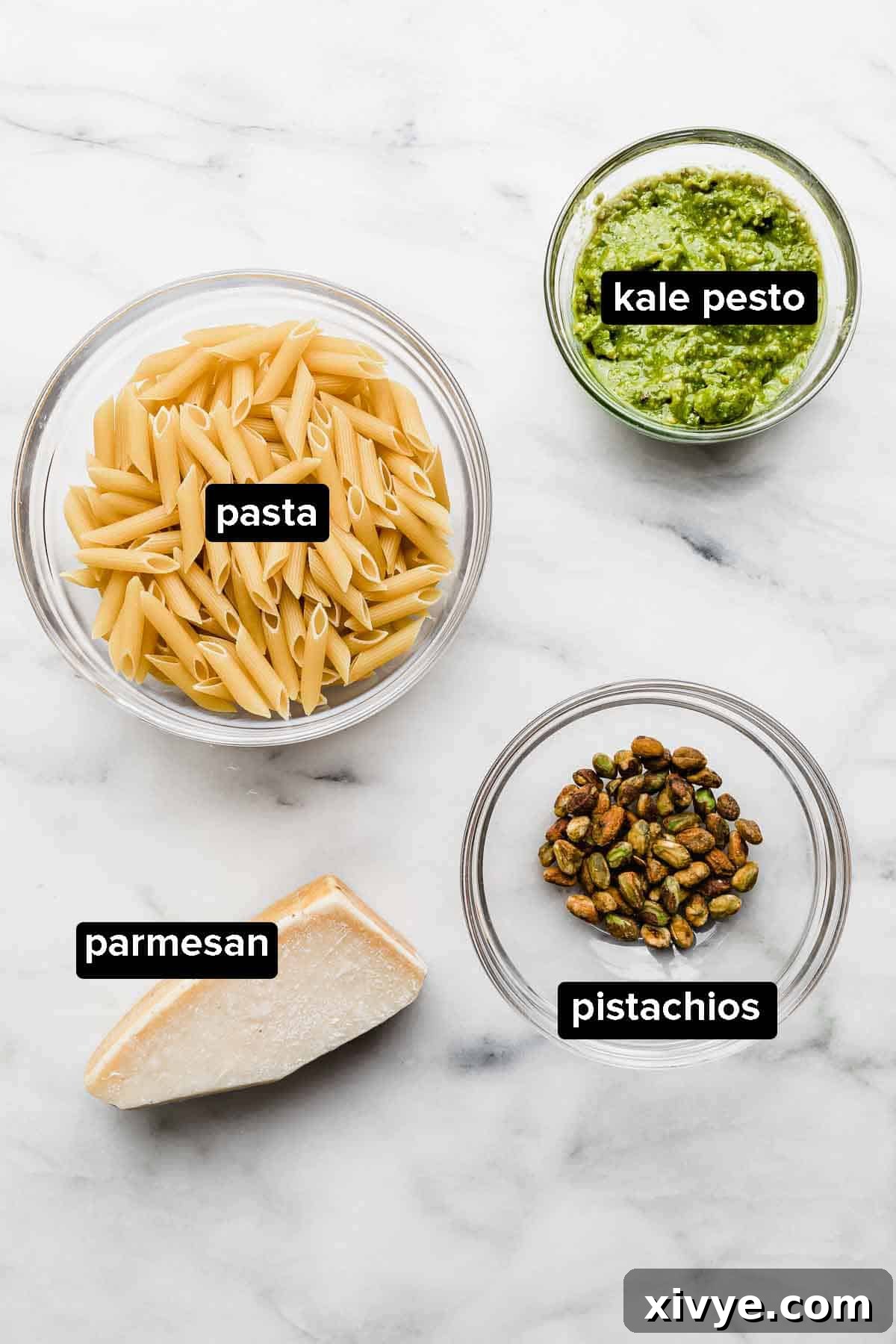 A vibrant flat lay of Kale Pesto Pasta ingredients on a white background, including fresh kale pesto in a bowl, uncooked pasta noodles, whole pistachios, and a block of parmesan cheese.