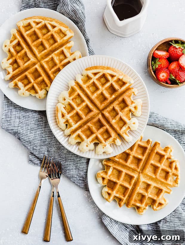 Wednesday's Little Indulgence 2 Three golden brown buttermilk waffles on separate white plates, artfully drizzled with maple syrup. A small white pitcher of syrup and a bowl of fresh, vibrant strawberries are visible off to the side, creating a tempting and wholesome breakfast scene, inviting viewers to indulge.