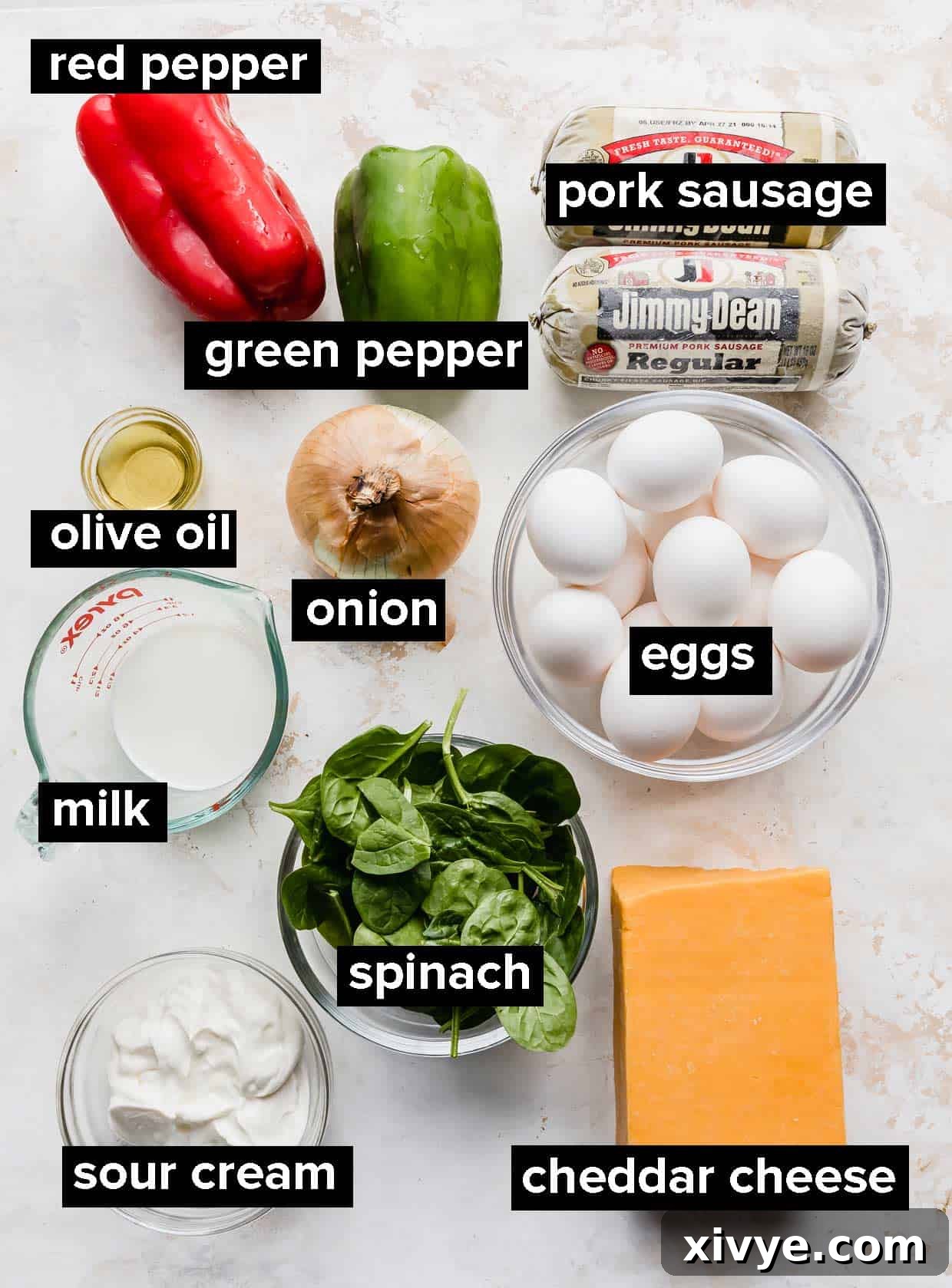 Simple Breakfast Bake 3 Various fresh ingredients including eggs, sour cream, milk, ground sausage, bell peppers, spinach, and shredded cheese, arranged on a textured white background, ready for making breakfast casserole.