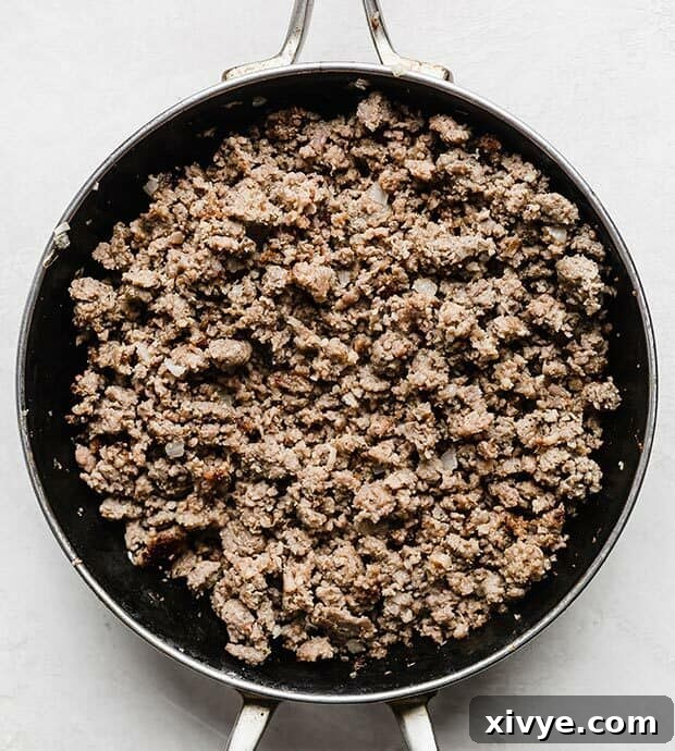 Simple Breakfast Bake 5 Overhead view of browned ground sausage and sautéed onions in a cast iron skillet, with a wooden spoon breaking up the meat.