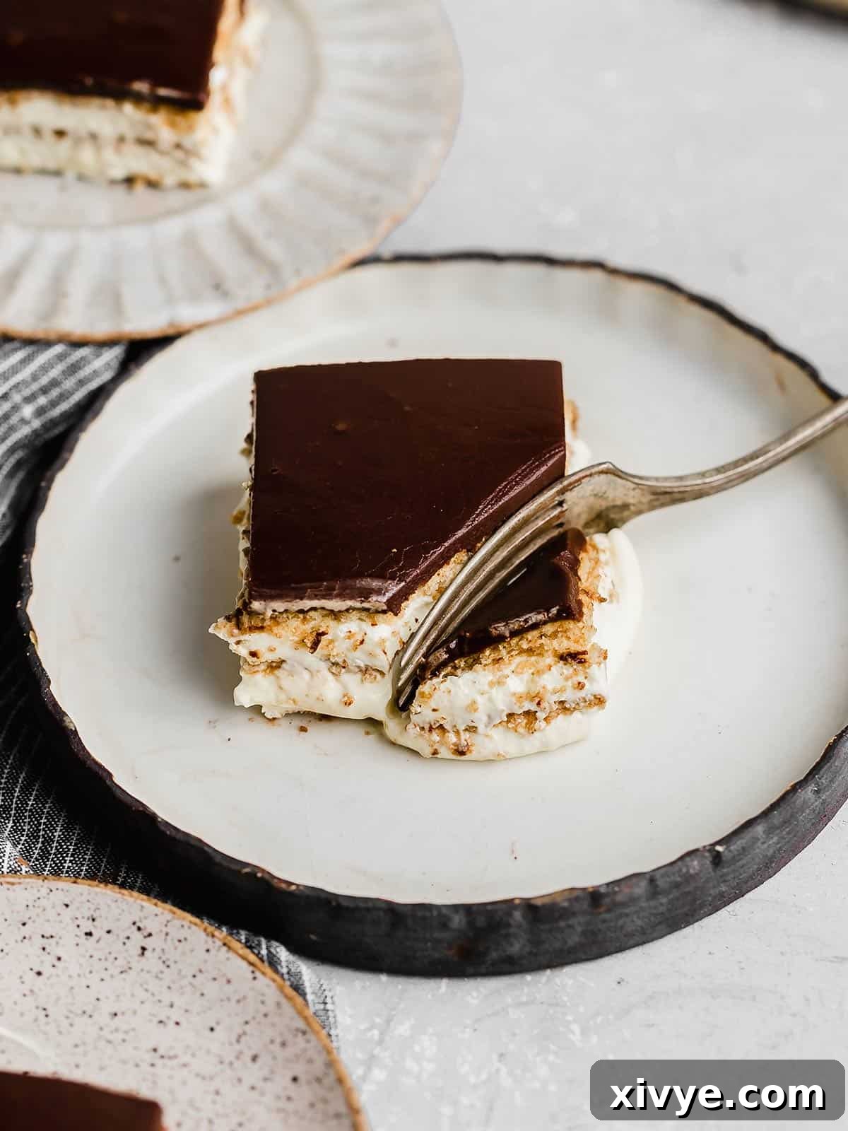 A square slice of Chocolate Eclair Cake on a black rimmed white plate, showcasing its beautiful layers and rich chocolate topping.