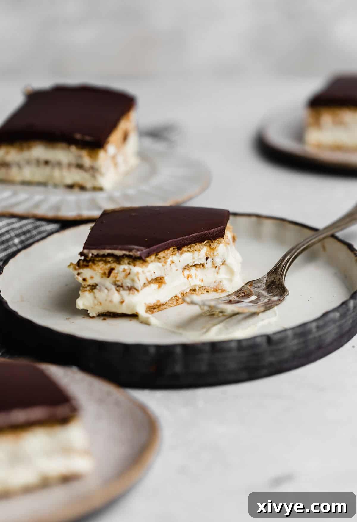 A no-bake Chocolate Eclair Cake, showcasing its layered structure of graham crackers and instant pudding topped with a rich chocolate glaze, presented on a white plate.