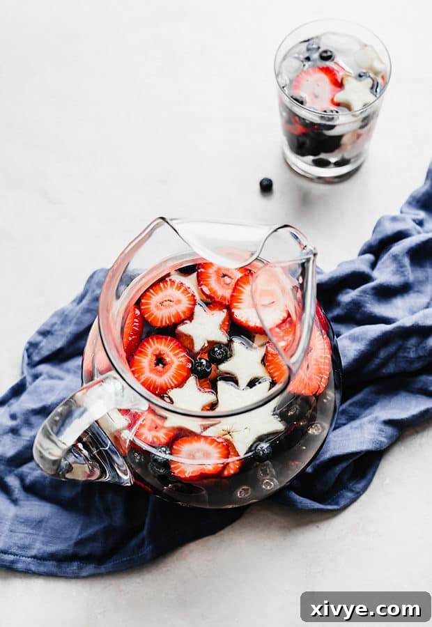 A large glass pitcher filled with vibrant red strawberries, deep blue blueberries, dark blackberries, and pristine white apple stars, ready for a 4th of July celebration.