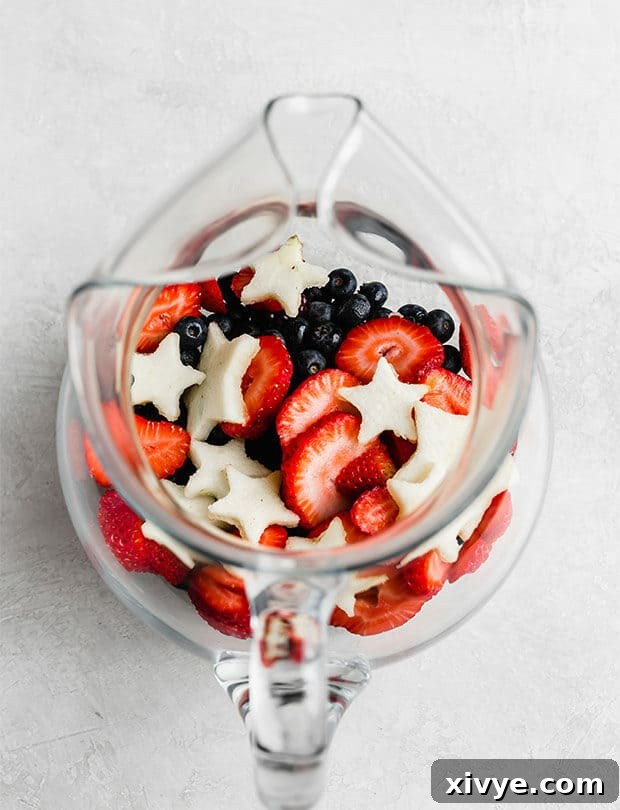 A beautifully arranged glass pitcher of 4th of July fruit-infused water, filled with red strawberries, white apple stars, and blue blueberries, set against a pristine white background.