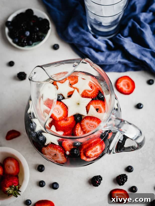 A close-up of a glass pitcher brimming with 4th of July fruit-infused water, showcasing an abundance of red strawberries, blue blueberries, dark blackberries, and delicate white apple stars.