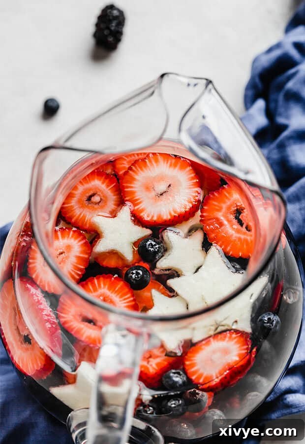 A close-up view of sliced strawberries, plump blueberries, and artfully cut star-shaped apples submerged in clear water within a glass pitcher, ready for a 4th of July gathering.