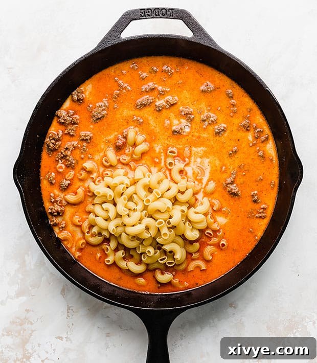 Ultimate Homemade Beef & Mac 6 An overhead shot of a cast iron skillet brimming with a rich red liquid, seasoned ground beef, and large macaroni noodles, simmering on the stove.