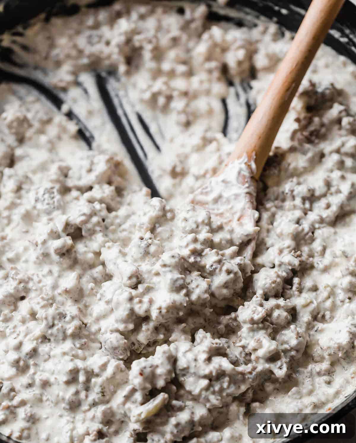 A creamy sausage dip in a black skillet and a wooden spoon scooping up some of the dip.