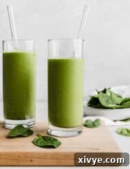 Two glasses full of a green protein smoothie on a wooden cutting board with a bowl of spinach in the background.