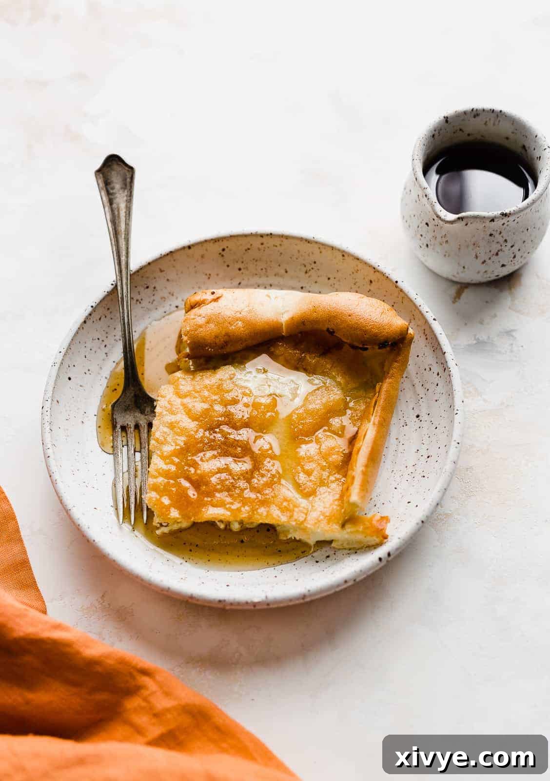 A square slice of German Pancake on a white plate with syrup overtop.