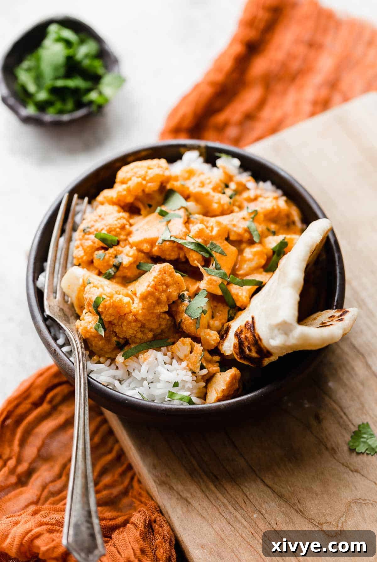 Red colored Indian Butter Cauliflower on a bed of white rice in a black bowl, with a piece of naan folded to the side and garnished with cilantro.