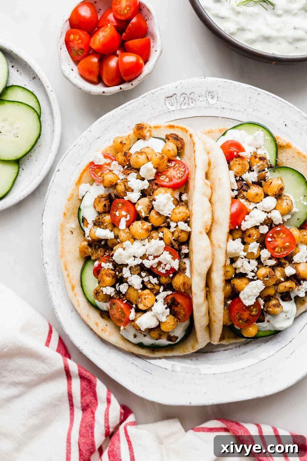 Mediterranean Chickpea Gyros 5 Two Chickpea Gyros on a white plate with a small bowl of cherry tomatoes, sliced cucumbers, and tzatziki sauce towards the top of the image.