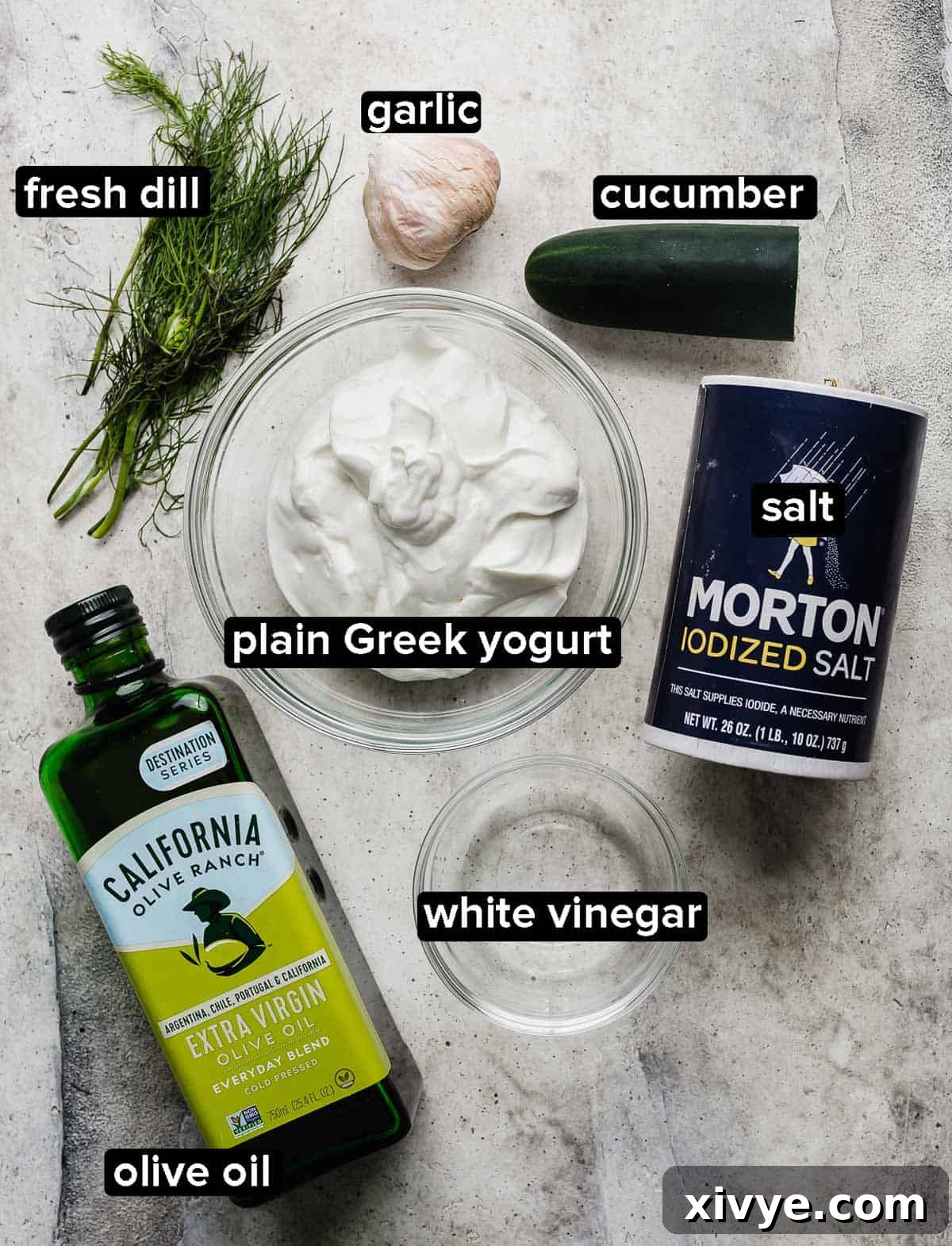 Tzatziki ingredients arranged beautifully on a light gray background: salt, a fresh cucumber, vibrant dill, golden olive oil, thick Greek yogurt, whole garlic cloves, and white vinegar.