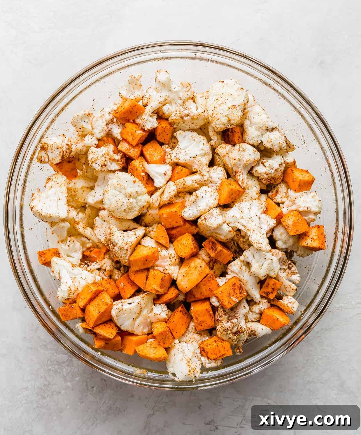 A glass bowl containing freshly chopped cauliflower and diced sweet potatoes, prepared for roasting.