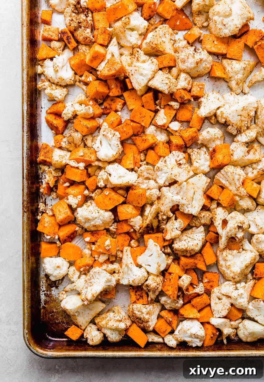 A baking sheet neatly arranged with olive-oil-coated and spiced cauliflower florets and diced sweet potatoes, ready for oven roasting.