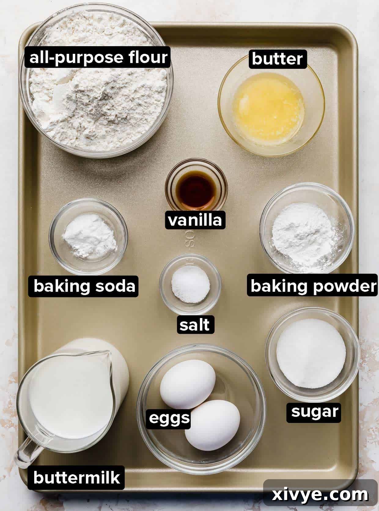 All the necessary ingredients for Homemade Buttermilk Pancakes, meticulously portioned into clear glass bowls and neatly arranged on an elegant bronze baking sheet, ready for mixing.