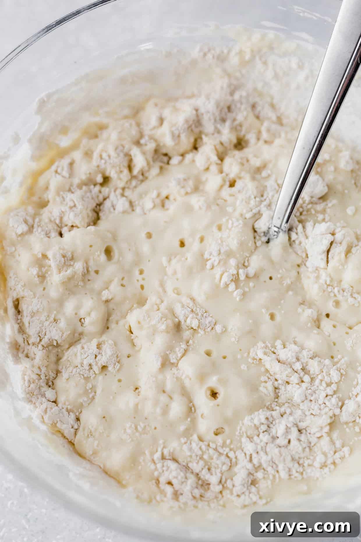 A fork is gently stirring the slightly lumpy, thick batter for Homemade Buttermilk Pancakes in a clear glass bowl, illustrating the ideal consistency before cooking.