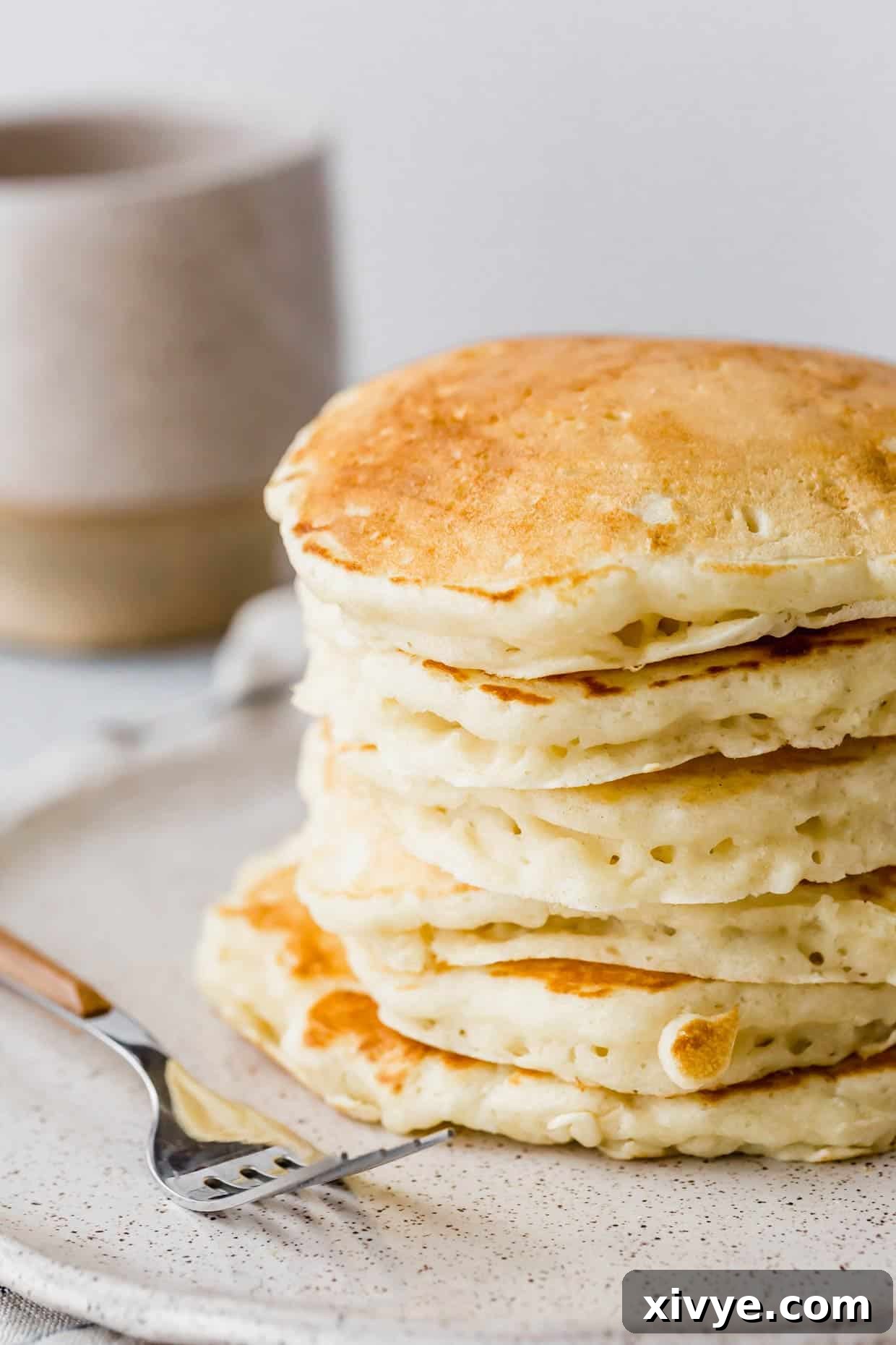 A tall, perfectly cooked stack of fluffy buttermilk pancakes rests on a simple white plate, ready to be enjoyed as a comforting breakfast.