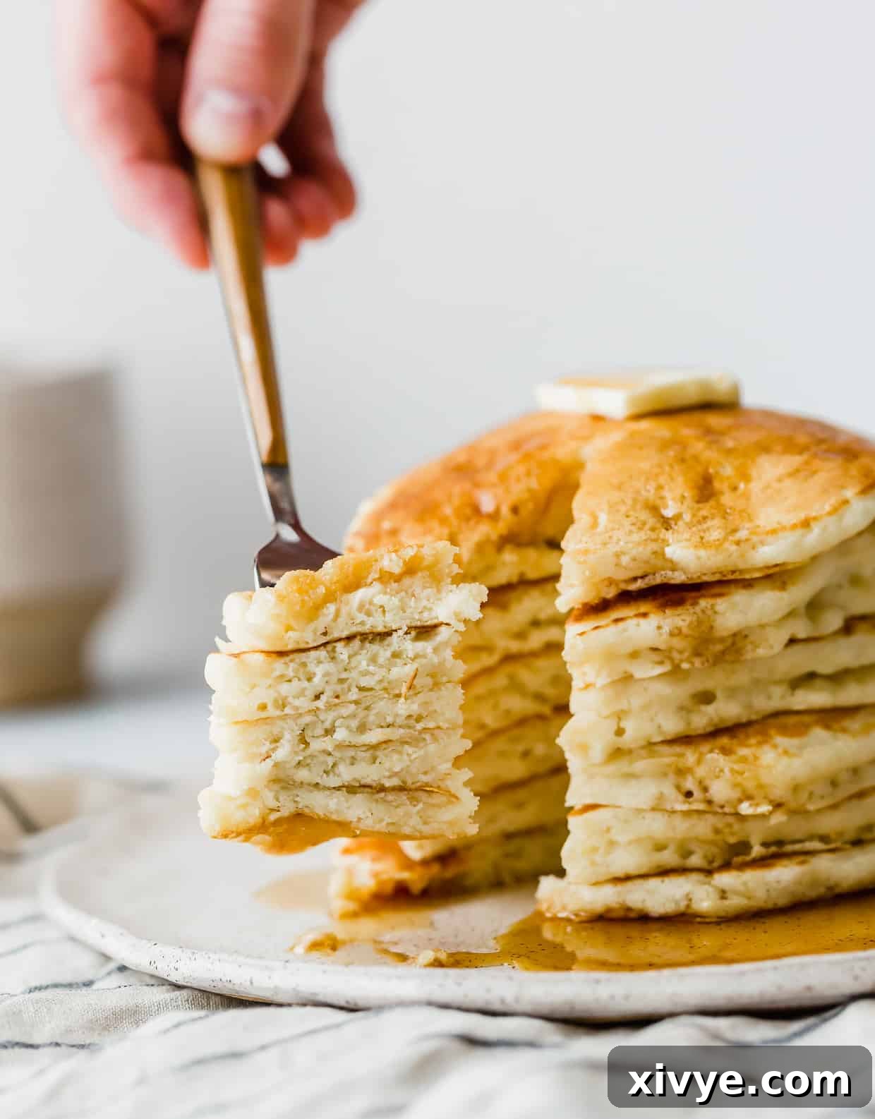 A fork holds a tantalizing bite of fluffy pancake, with a mouth-watering stack of golden buttermilk pancakes blurred in the background, inviting you to indulge.