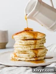 Golden maple syrup being generously poured over a towering stack of perfectly fluffy buttermilk pancakes, presented on a pristine white plate, promising a delightful breakfast.