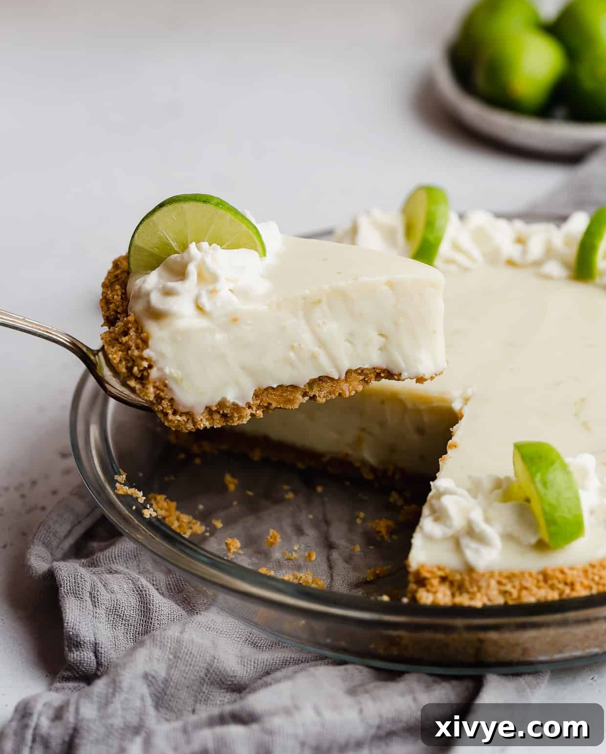 Velvet Key Lime Pie with a Sour Cream Kiss 2 A metal spatula holding up a slice of key lime pie with a golden brown crust, topped with light green whipped cream and a lime slice.
