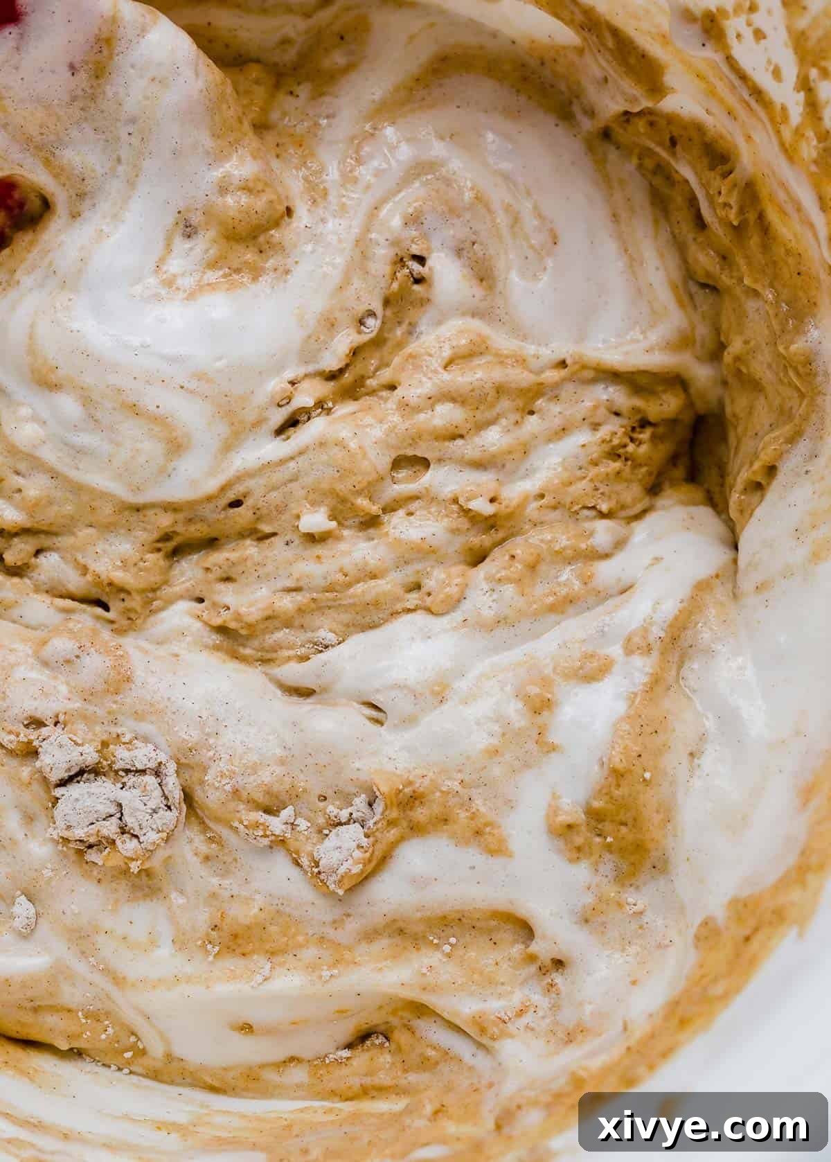 A close-up shot of fluffy whipped egg whites being gently folded into a rich, orange pumpkin waffle batter with a spatula, creating a marbled effect and highlighting the delicate mixing process.