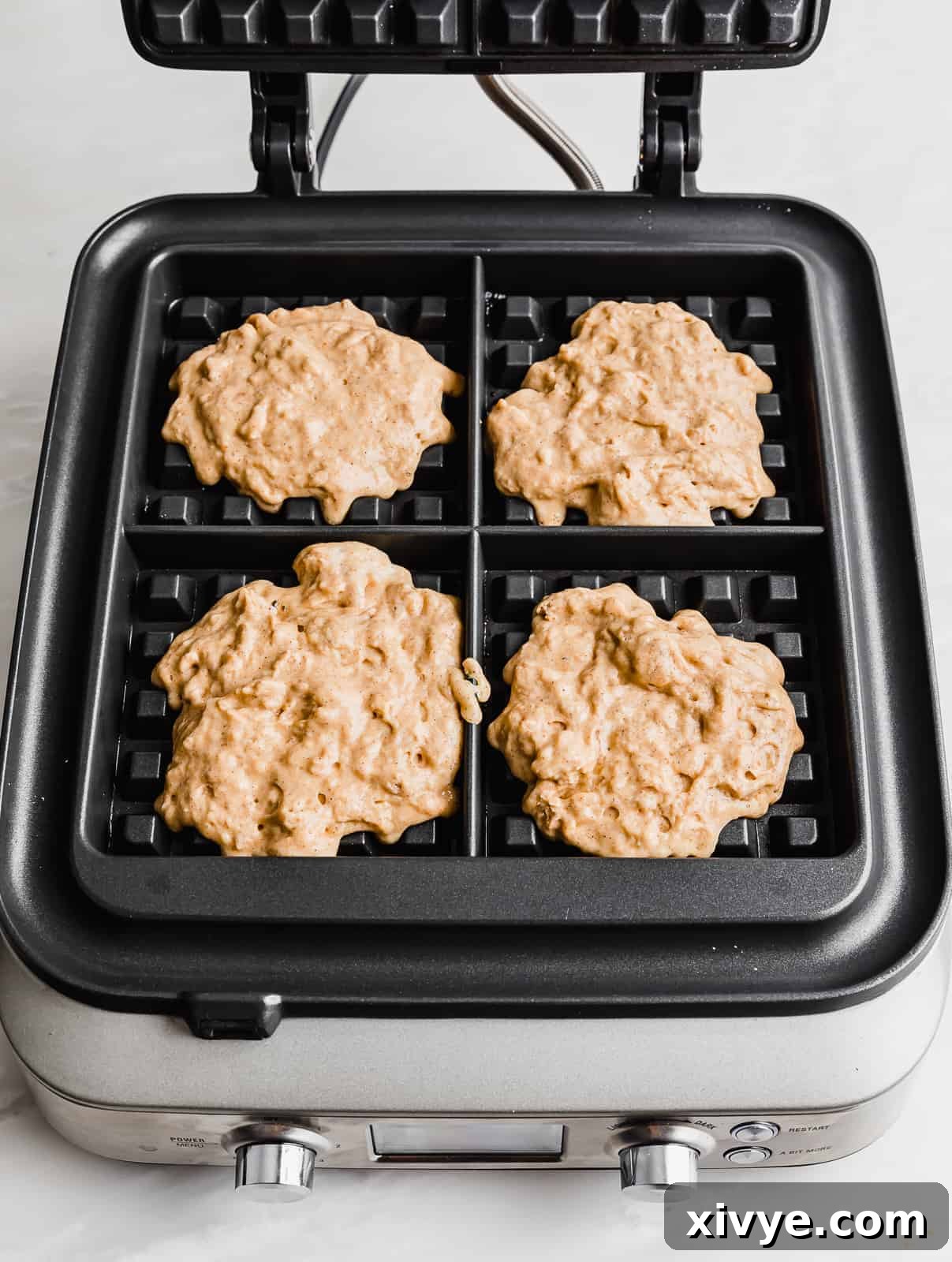 A square waffle iron containing pumpkin waffle batter spread evenly into its four compartments, showing the textured grid ready for cooking.