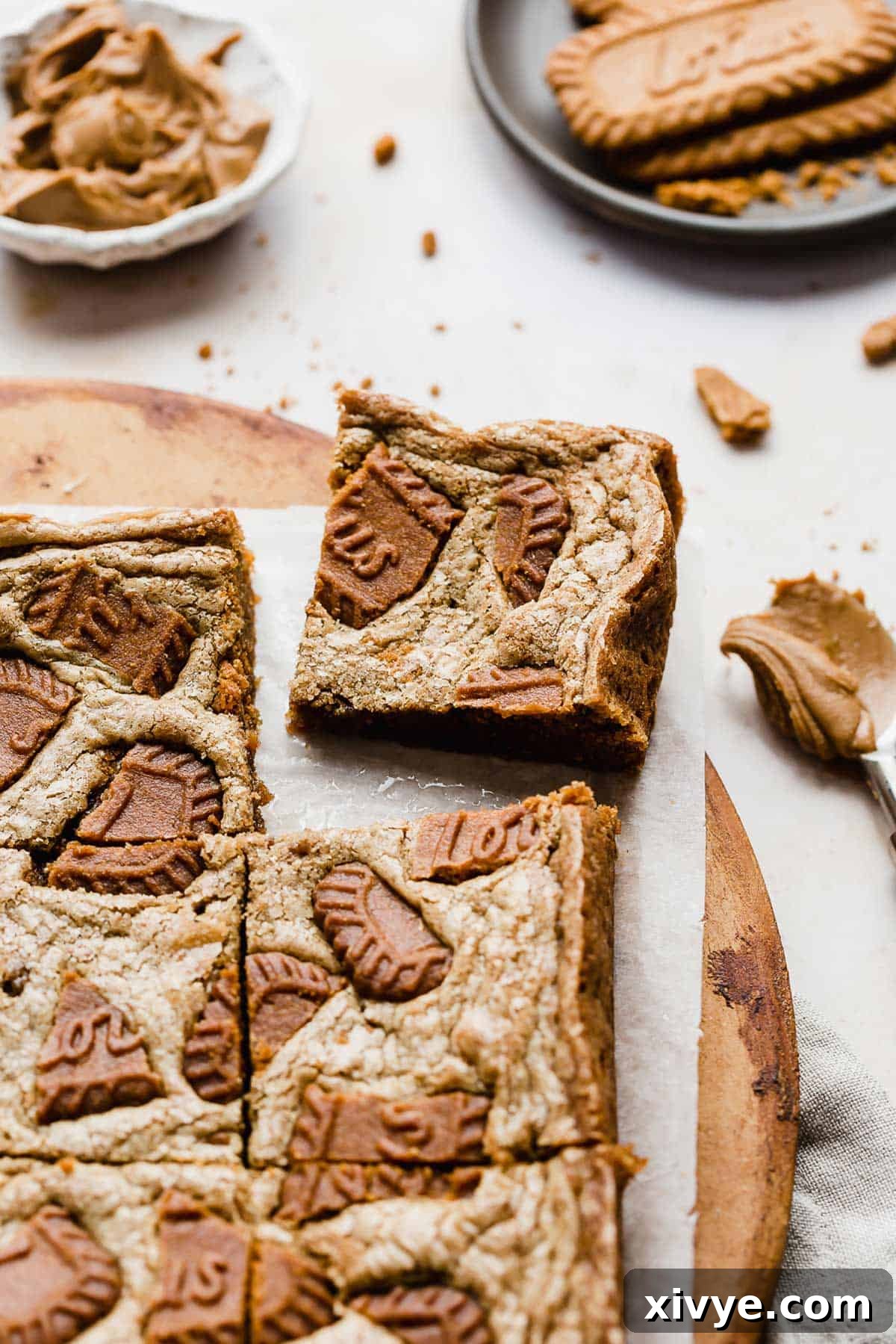 A square of Biscoff Brownies on a white parchment paper with Biscoff cookies in the background.