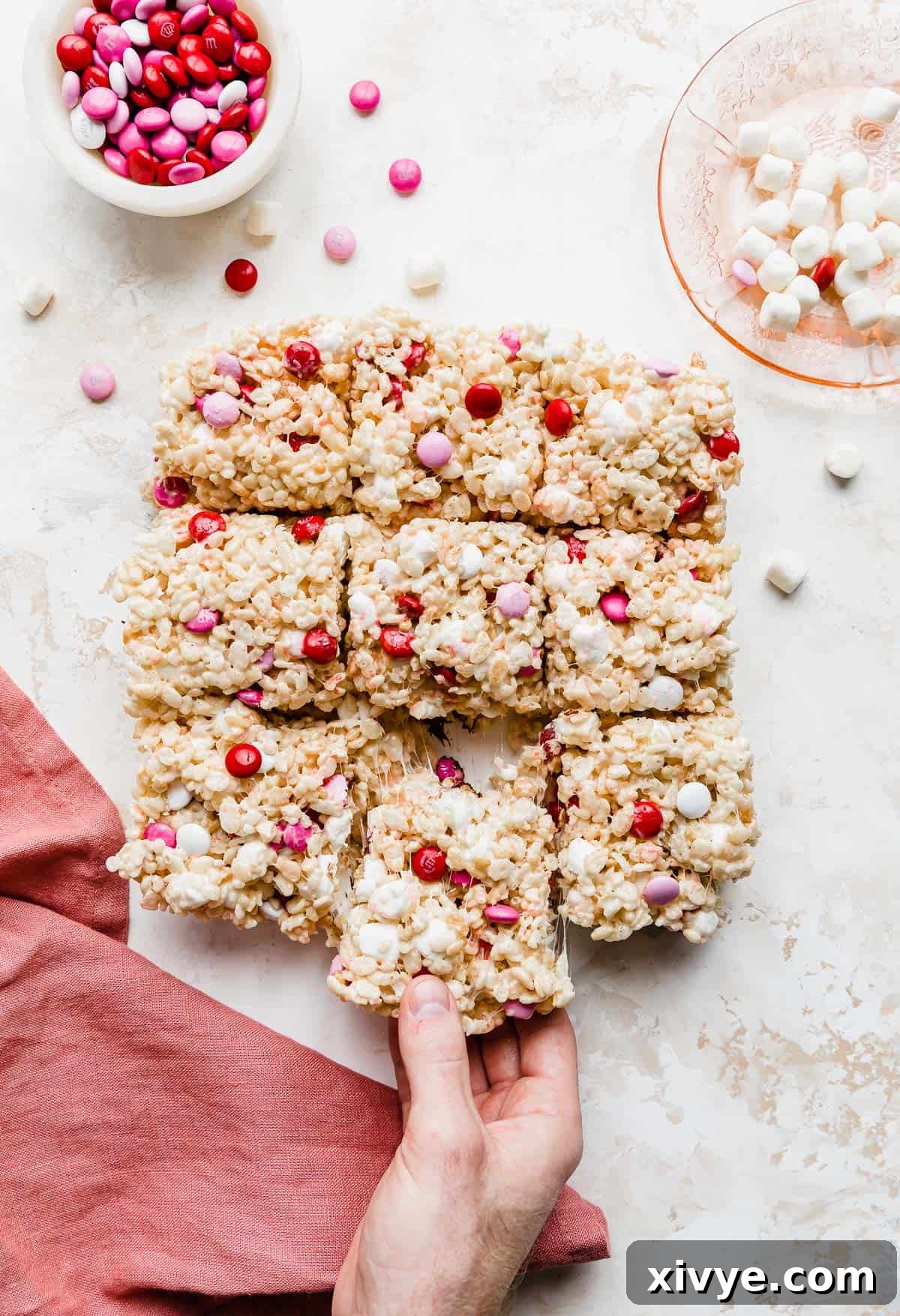 Colorful M&M Crispy Bars 2 A hand grabbing a M&M Valentine Rice Krispie Treat showing the gooey marshmallow pulling, emphasizing its soft and chewy texture.