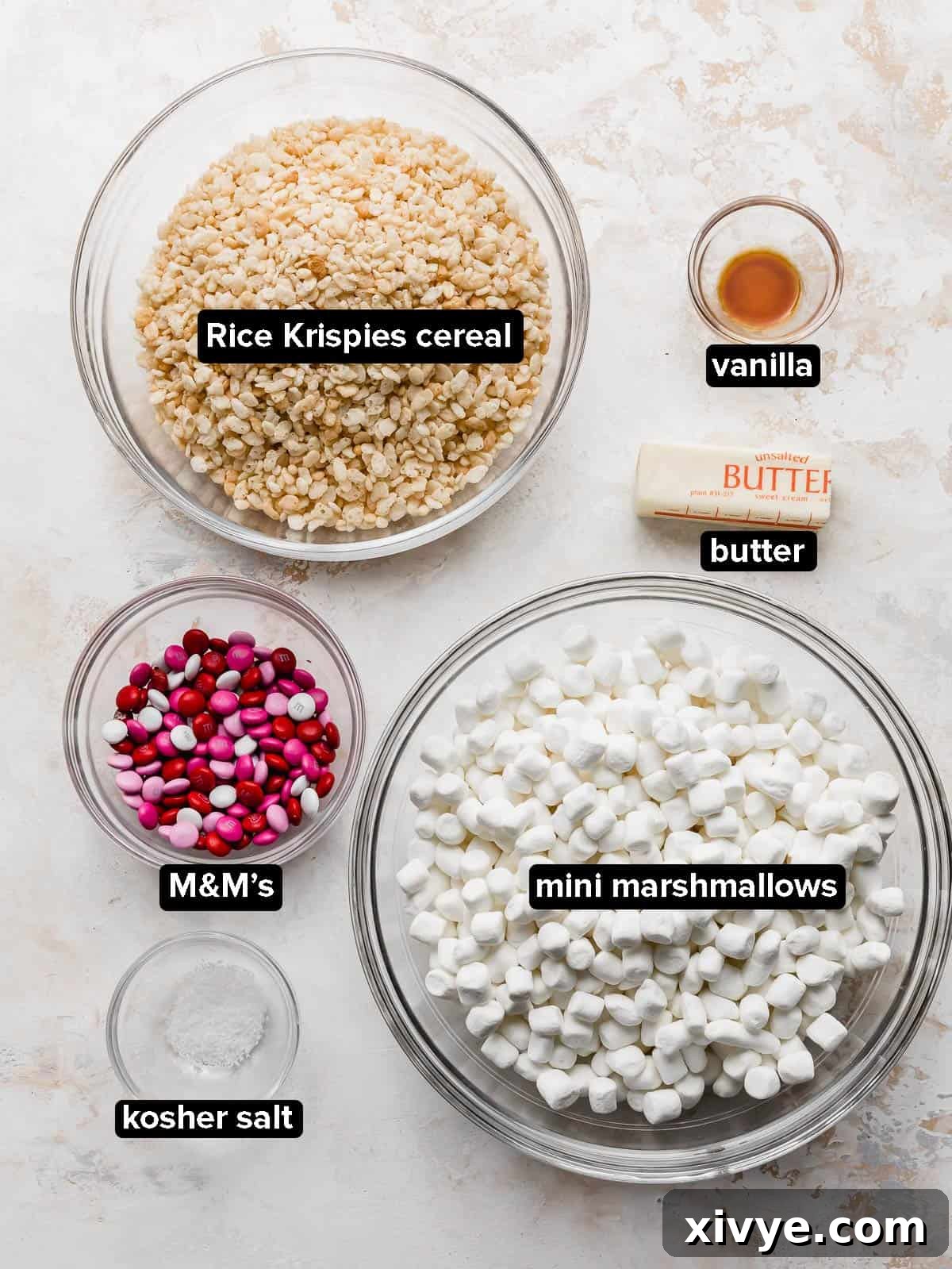 Colorful M&M Crispy Bars 3 Valentine Rice Krispie Treats ingredients in a glass bowl on a white background, including butter, marshmallows, Rice Krispies, salt, vanilla, and M&Ms.