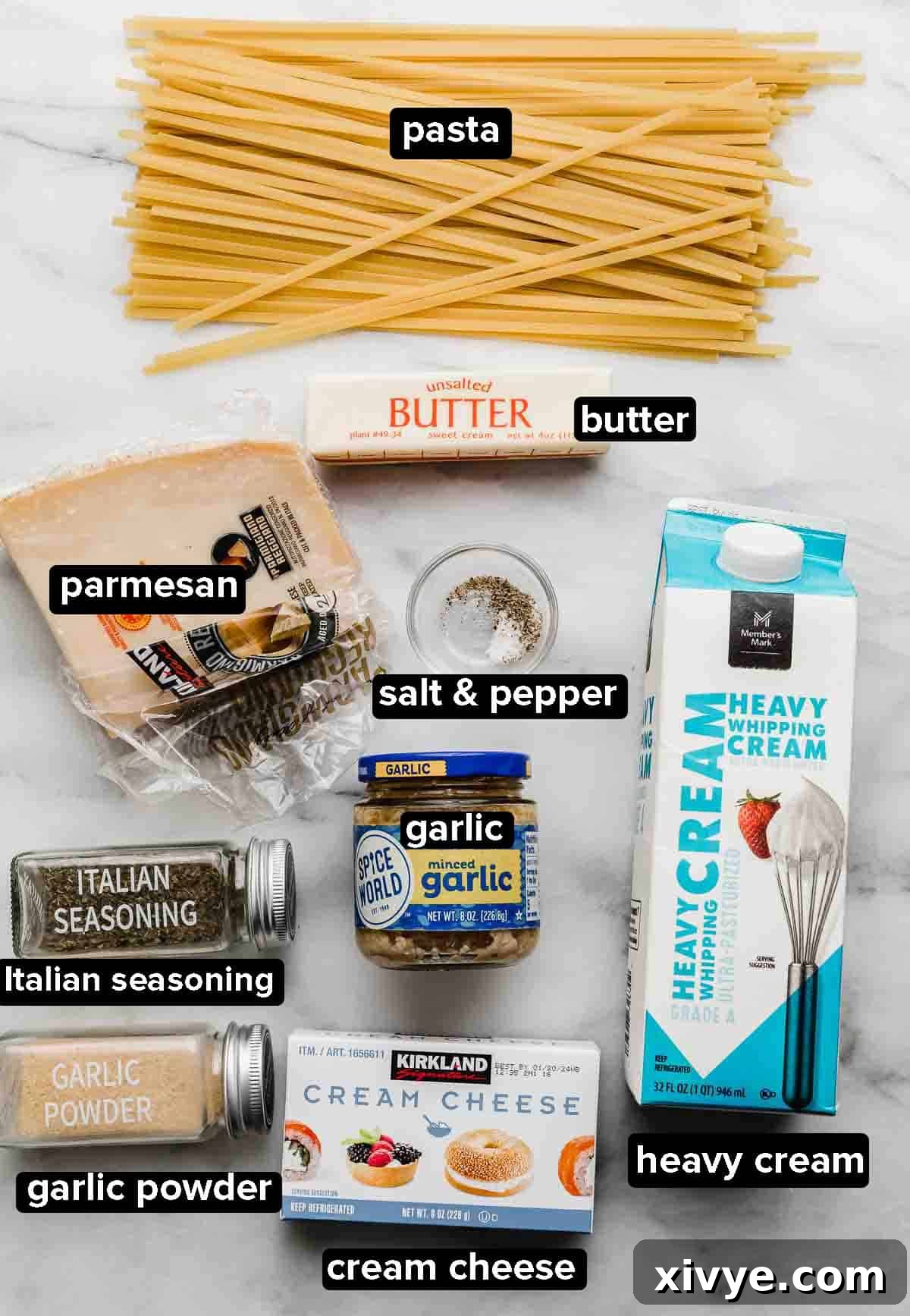 Fettuccine Alfredo ingredients on a white background: heavy cream, cream cheese, parmesan, pasta, salt and pepper, garlic, and Italian seasoning laid out for cooking.