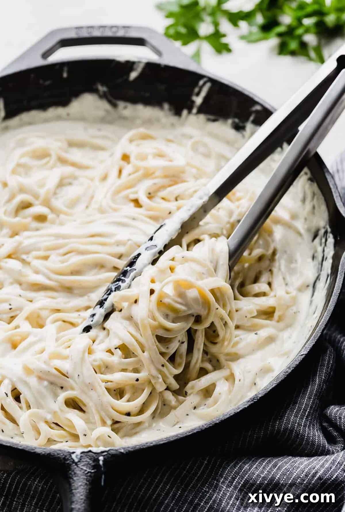 Kitchen tongs stirring creamy Fettuccine Alfredo in a black skillet, showing the rich texture.