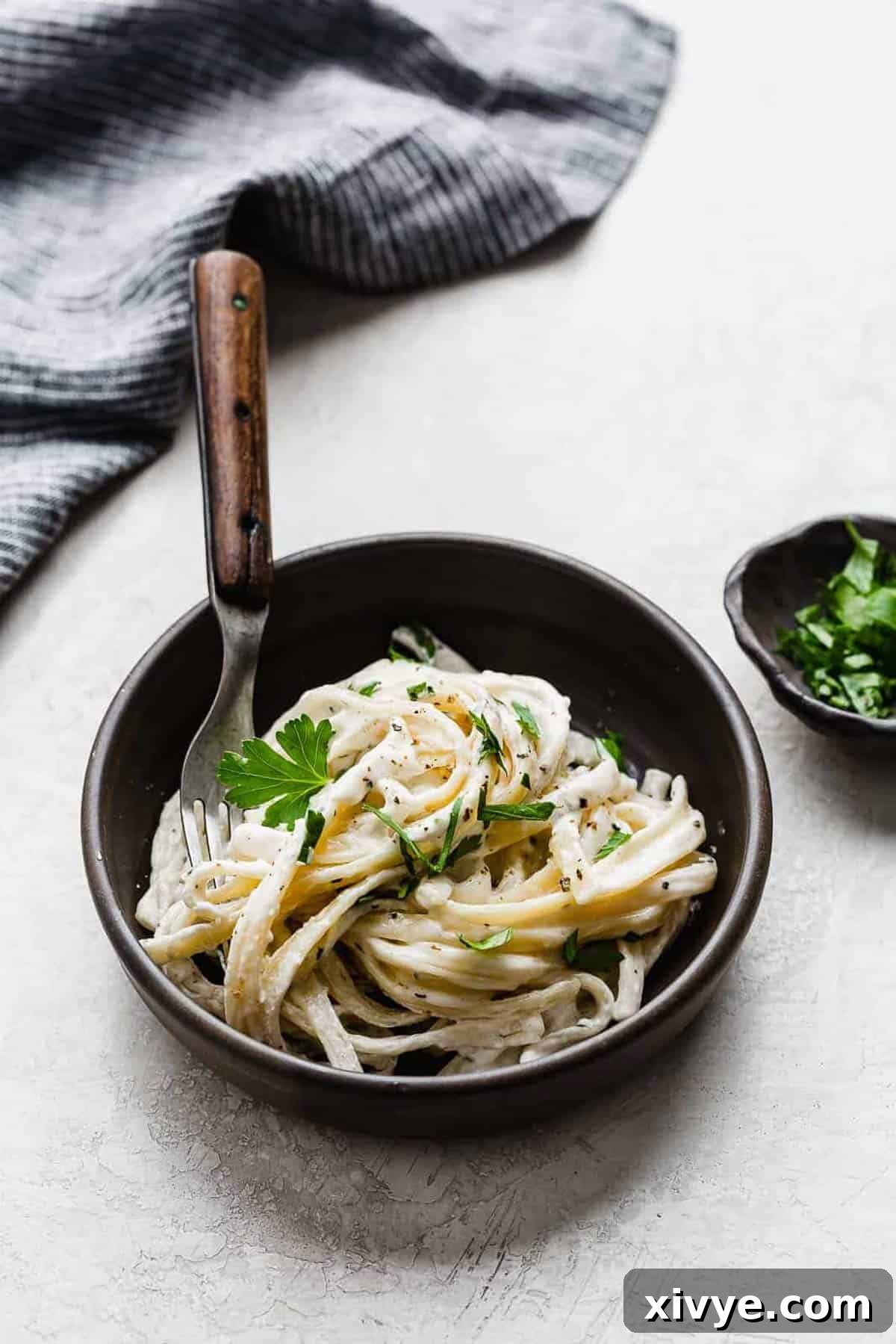 Fettuccine Alfredo in a black bowl on a gray background, garnished with fresh parsley.
