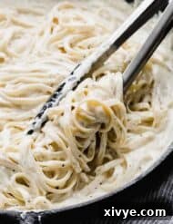 Creamy Fettuccine Alfredo in a black skillet with kitchen tongs being used to mix the creamy pasta.