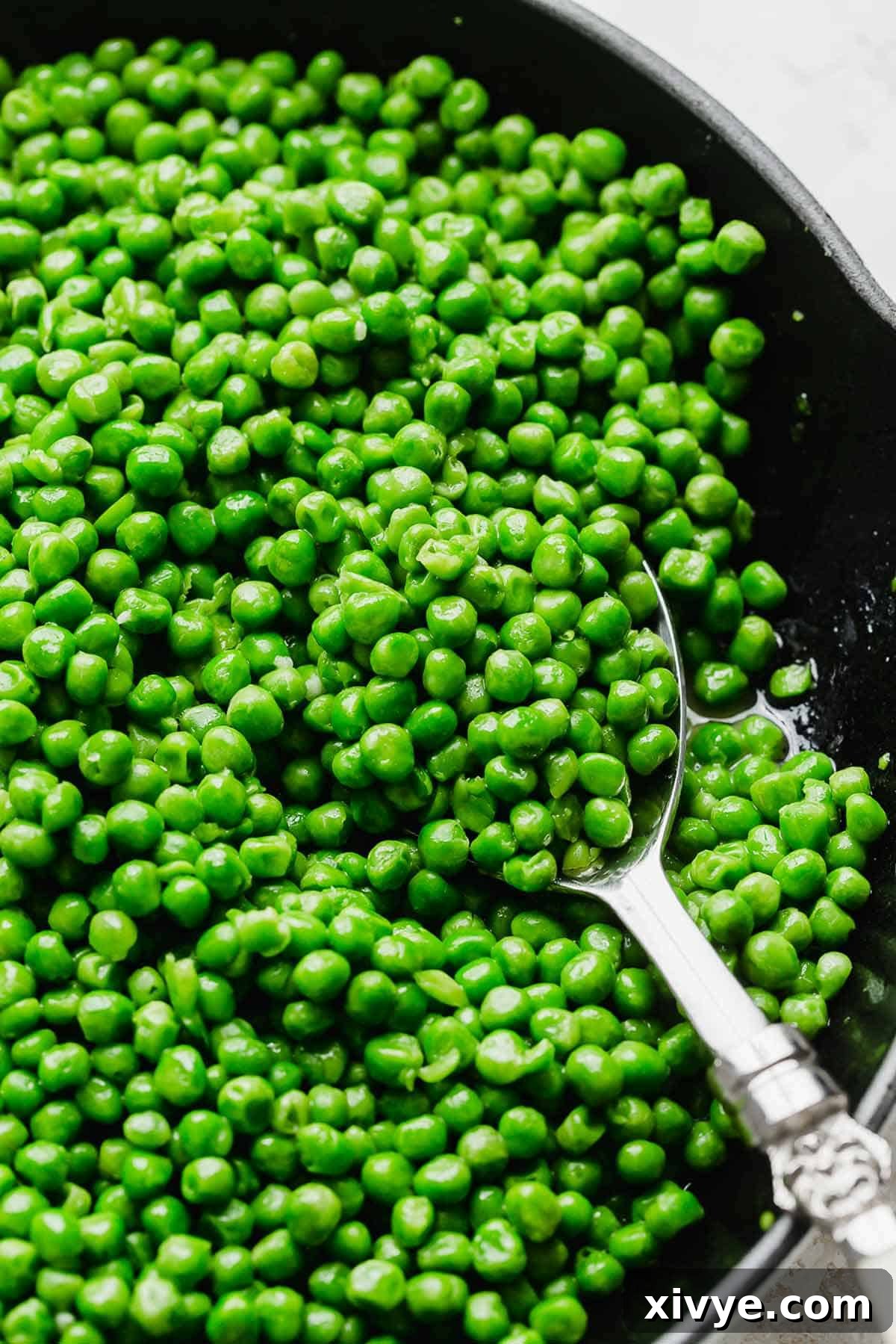 Elevate Your Frozen Peas 2 A silver serving spoon scooping up vibrant green, perfectly cooked peas from a cast iron skillet, showcasing the ideal texture achieved by sautéing.